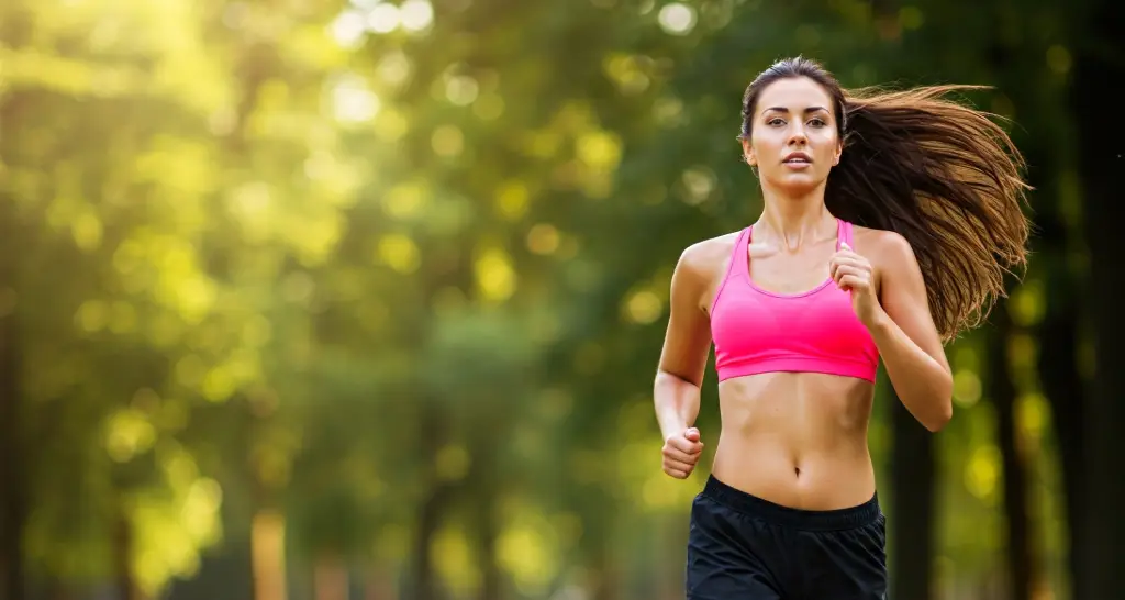 Una mujer joven y sonriente hace footing en un parque soleado. Su movimiento fluido y su expresión relajada reflejan el bienestar físico y mental que el ejercicio aporta, similar a cómo la aromaterapia puede mejorar el estado de ánimo y reducir el estrés.